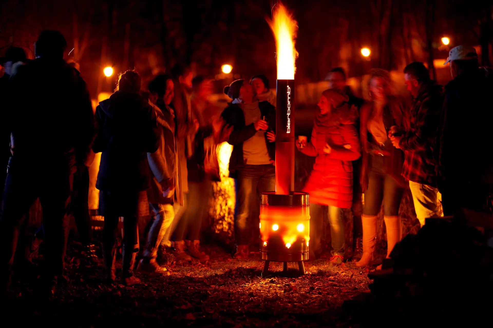 Brennende bierbomb-Feuertonne bei Nacht, eine Flamme schlägt aus dem Schornstein, Leute stehen im warmen Licht drumherum