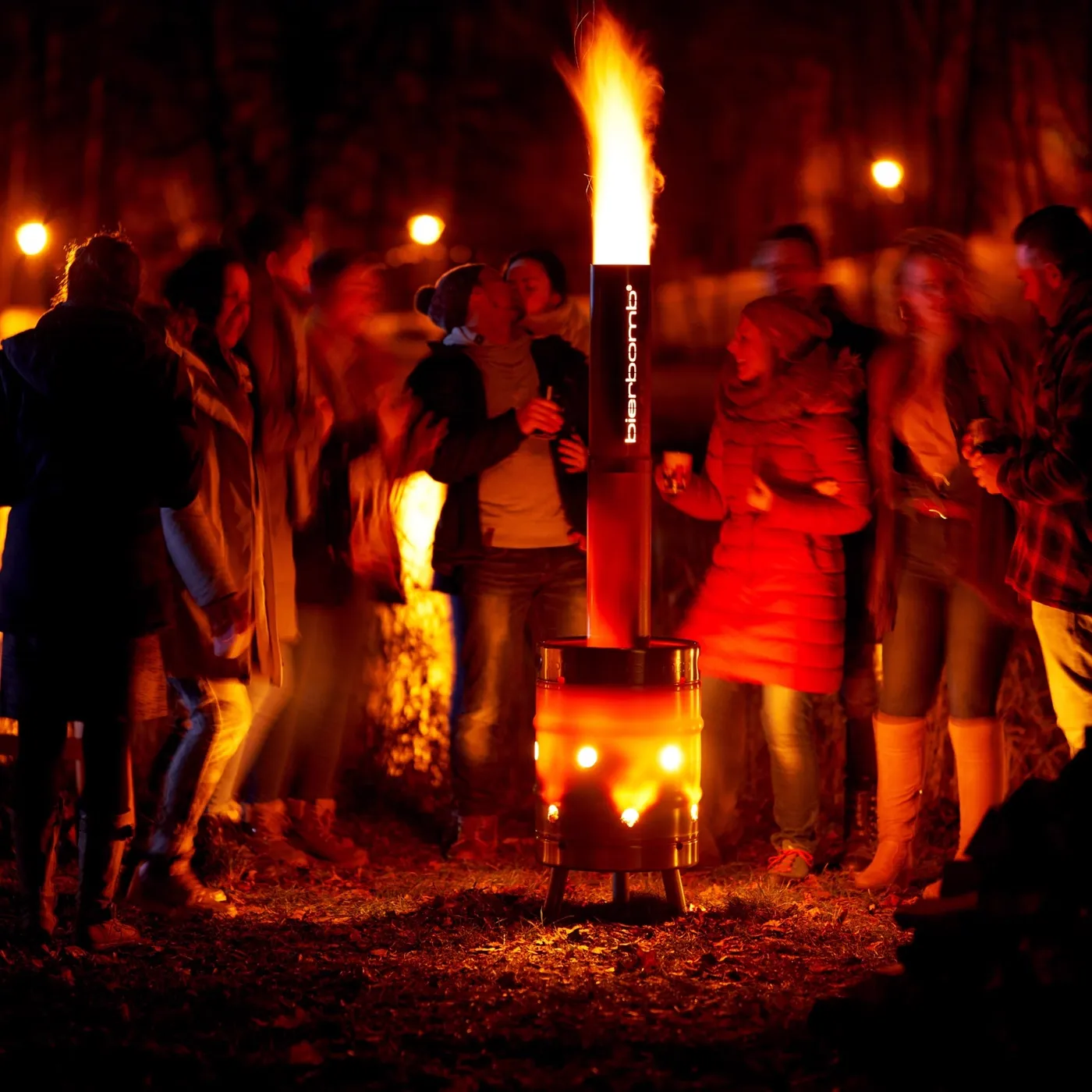 Gruppe von Gästen steht bei Nacht um die brennende bierbomb® Feuertonne, Flamme über dem Schornstein, die Luftlöcher glühen orange